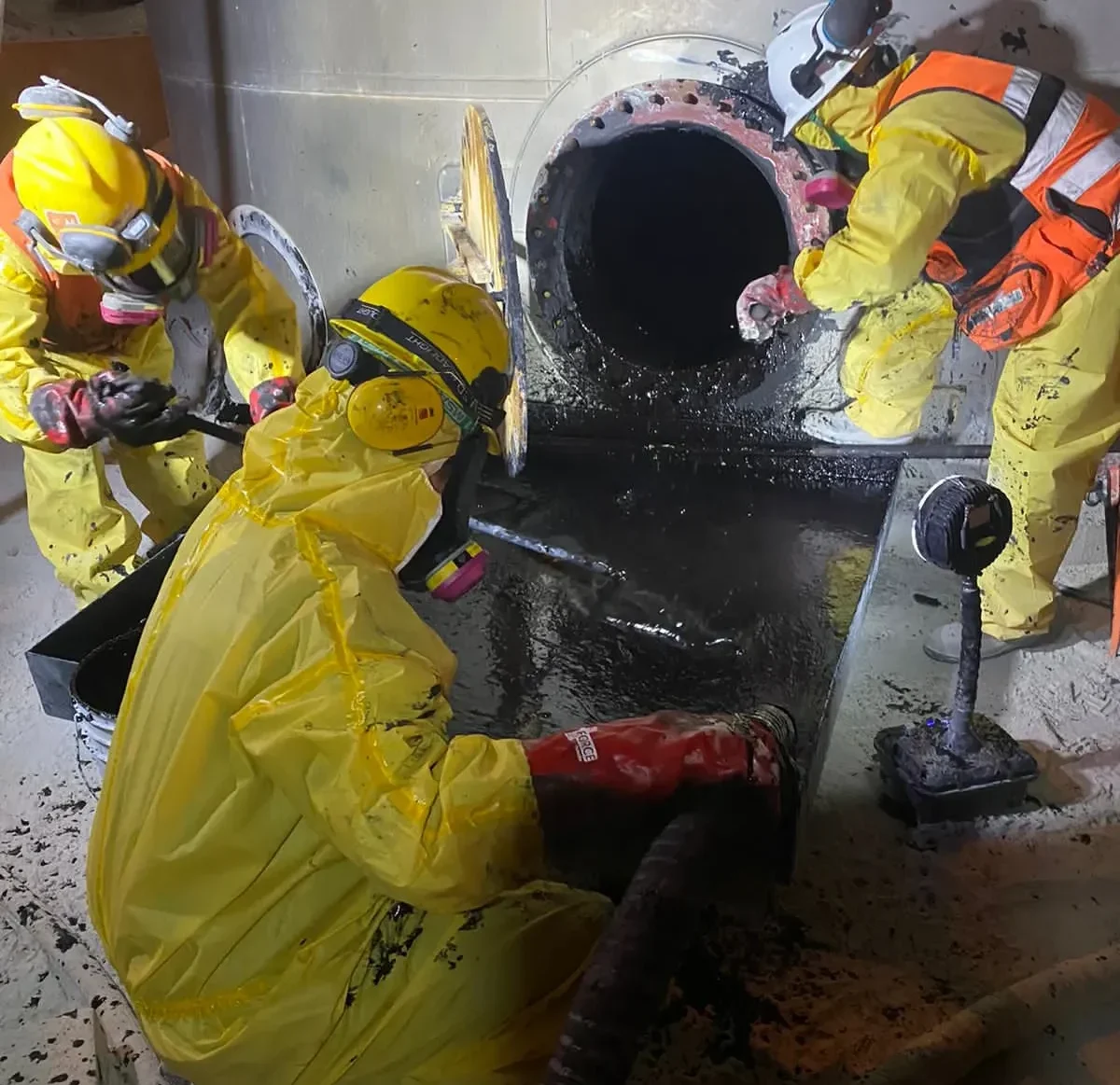 Trabajadores con trajes de protección química realizando tareas de descontaminación industrial en un espacio confinado dentro de una instalación de procesos.