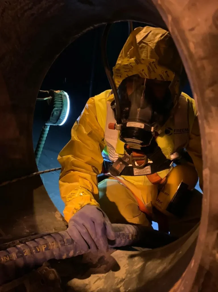 Trabajador con traje de protección química y equipo de respiración realizando limpieza e inspección dentro de un estanque industrial.